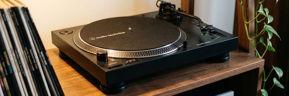 Audio-Technica AT-LP120XUSB direct-drive turntable on a walnut hi-fi shelf beside a row of vinyl records and a trailing plant